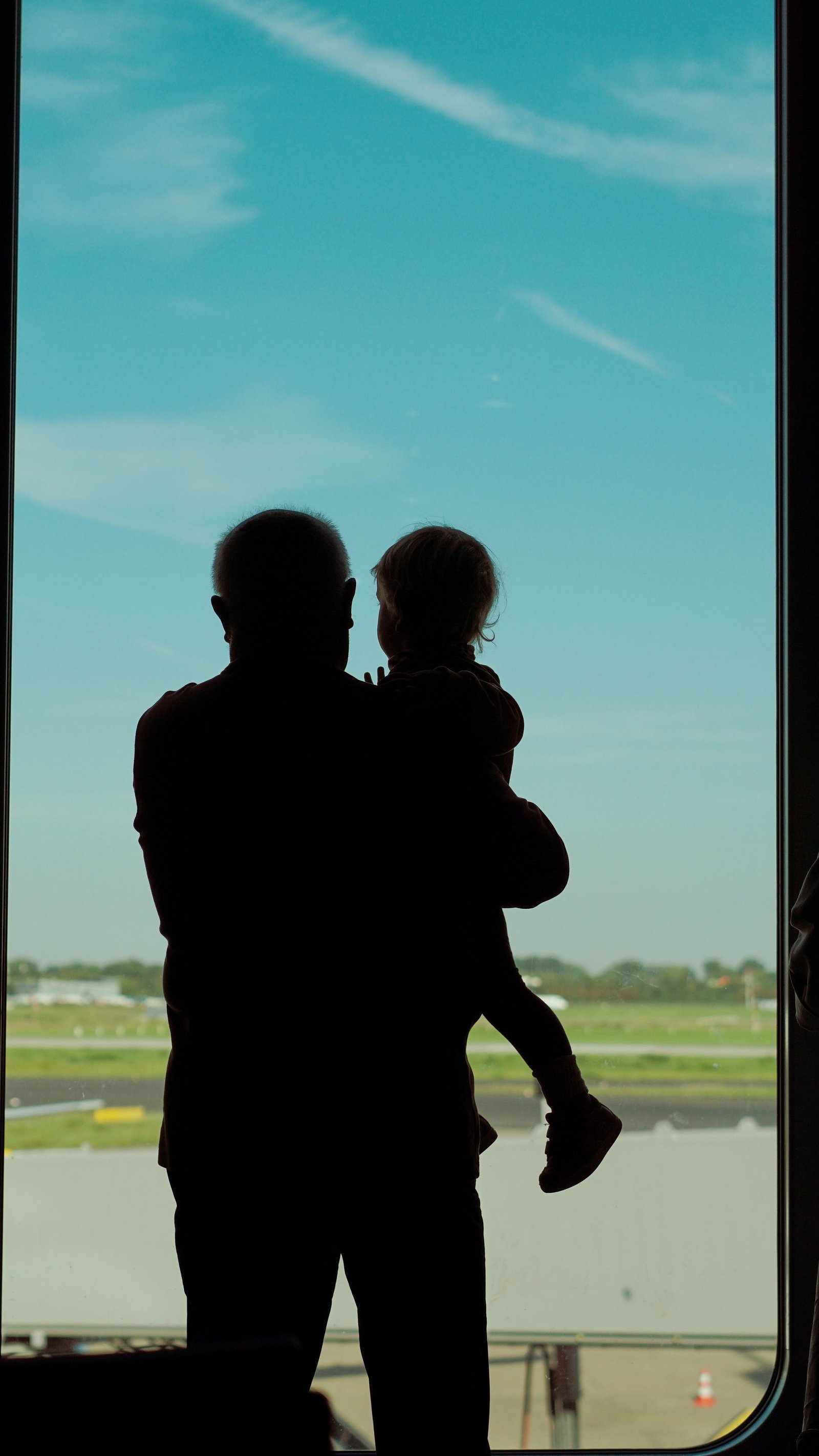 A parent holding a child at a wide airport window with open sky beyond