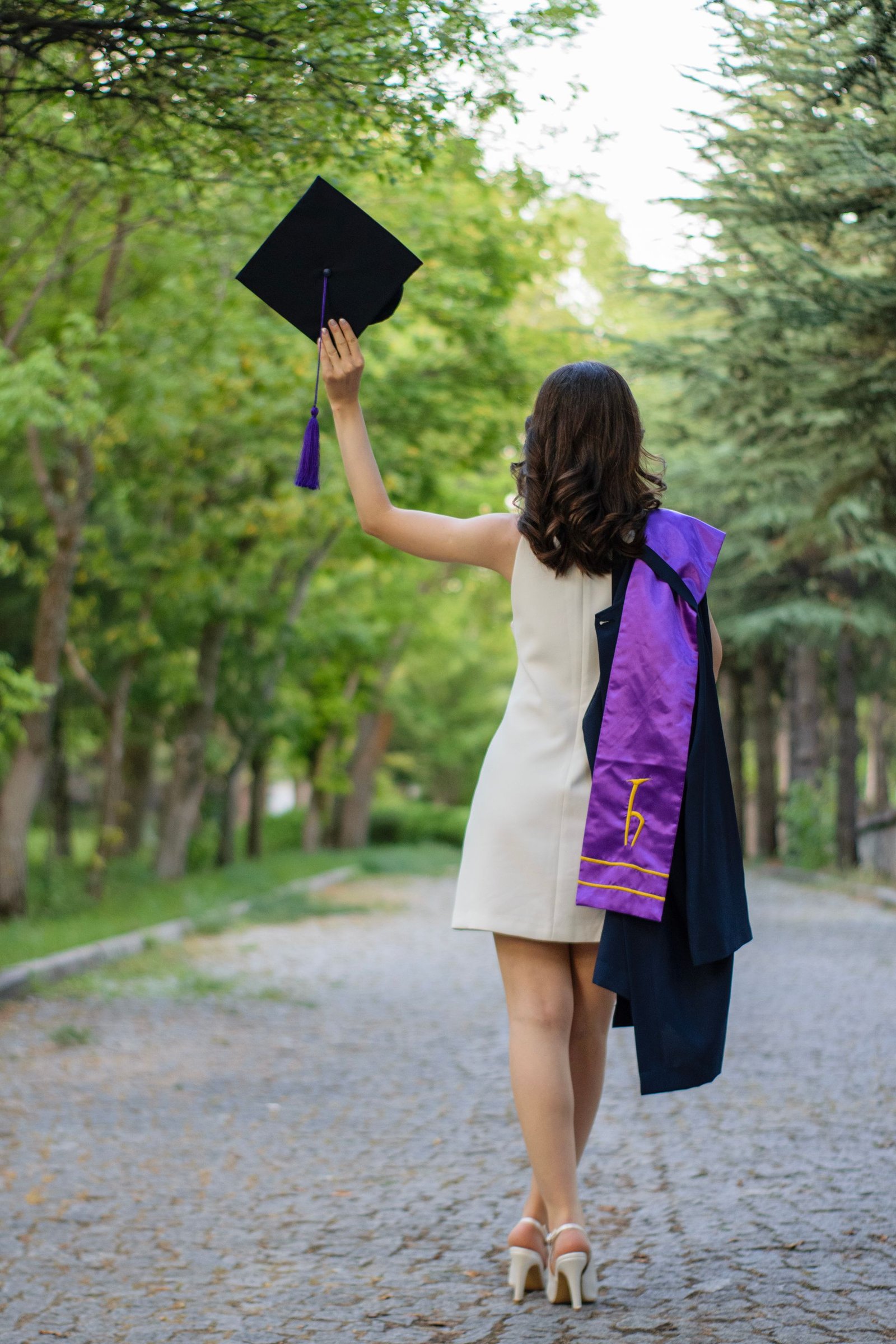 A graduate in a gown raising her cap on a tree-lined park path