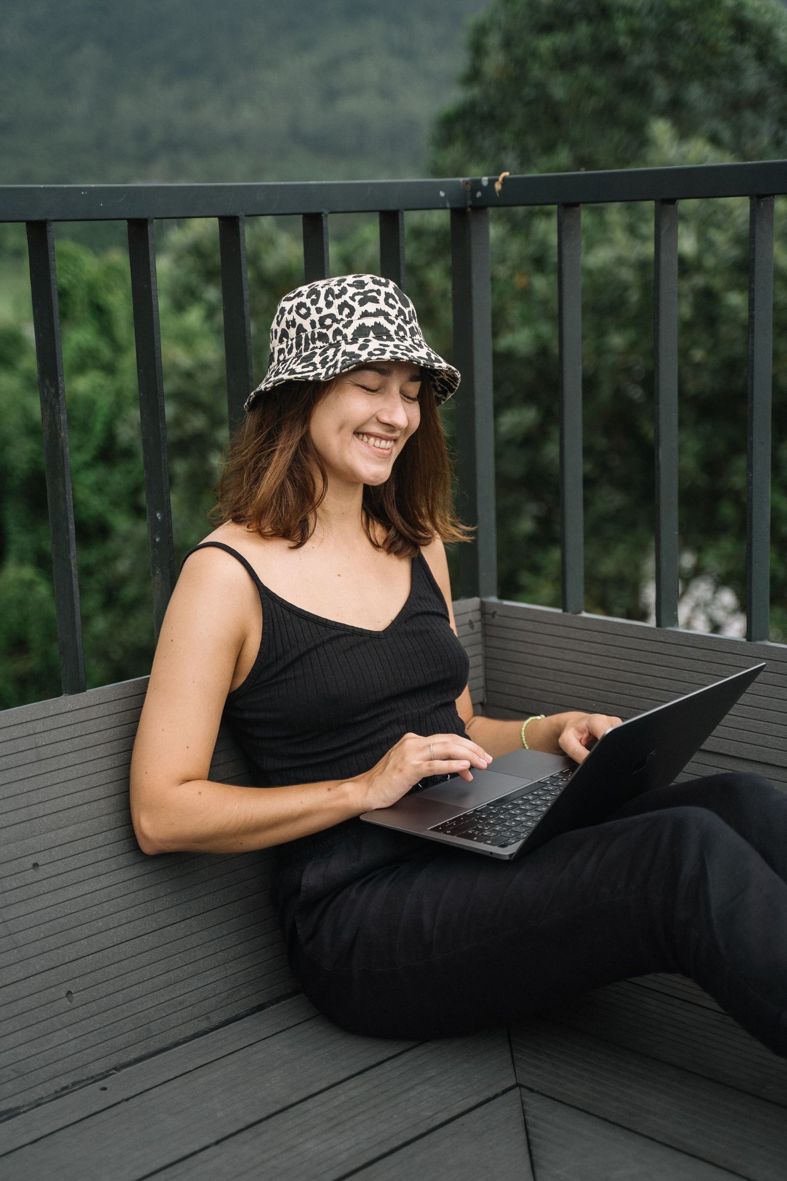 A smiling woman working on a laptop on a balcony surrounded by greenery