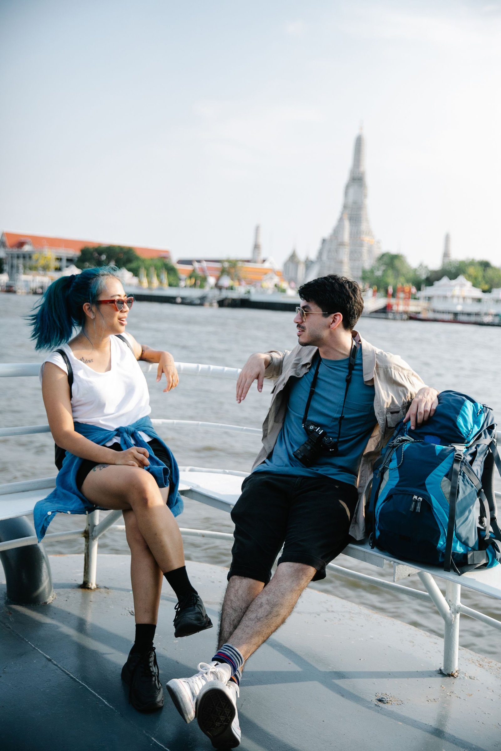 Two travelers relaxing on a ferry deck with their backpacks, a riverside temple behind them